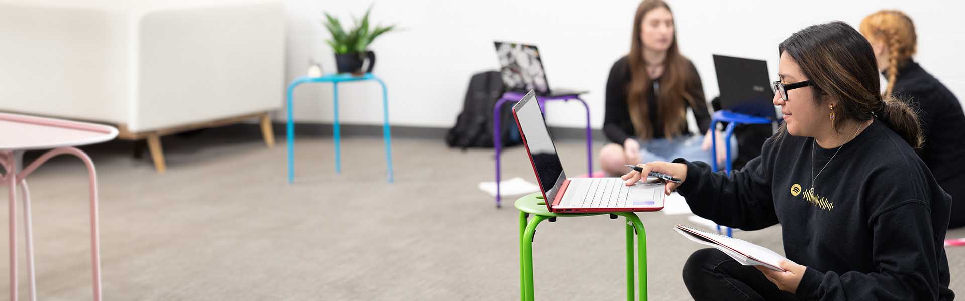 Female student sitting on the floor typing on laptop in front of her with other students in the background