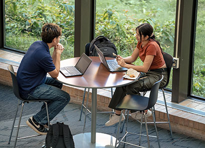 A male and female student studying in an atrium with big windows looking out at nature.