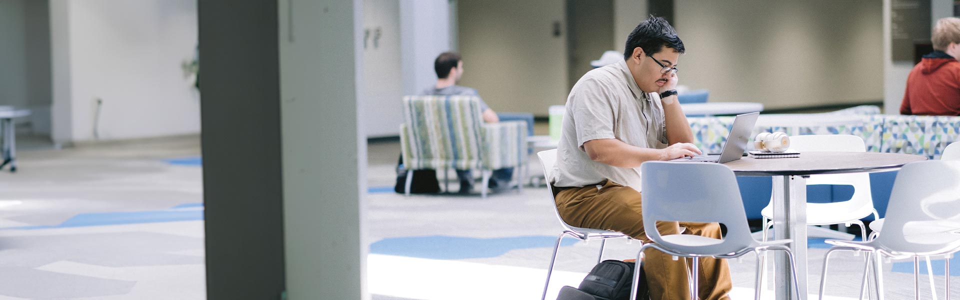 A student sitting in a common area at a table looking at their laptop